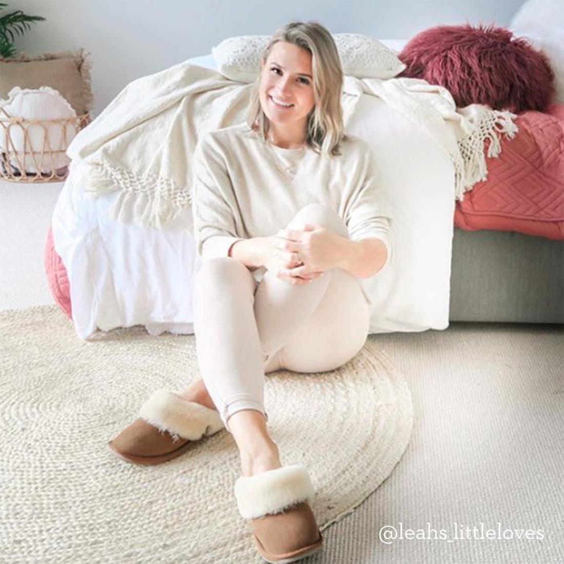 A cheerful woman relaxes at home in EMU NZ Eden Platinum Chestnut Scuff Slippers, with soft sheepskin, slip-on style, and sturdy sole.  ksnz