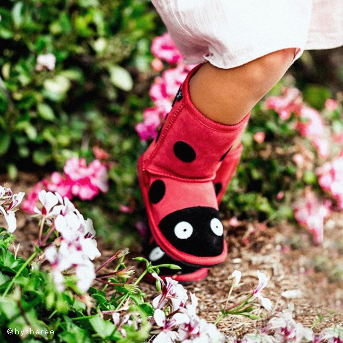 A child in pink pants wears EMU Kids Creatures Red Ladybird Wool Boots, known for comfort and durability, standing among vibrant pink and white flowers., 51426, 9330071131183, 9330071131190, 9330071131206, 9330071131213, 9330071131220, 9330071131237, 9330