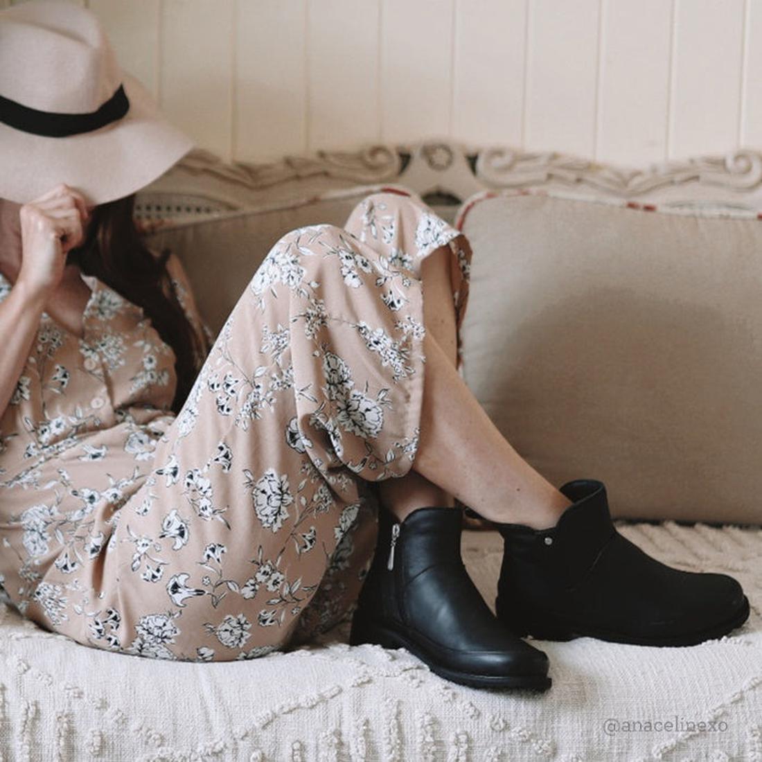 A woman in a floral beige jumpsuit and EMU Women's Nashira Ankle Boot Black sits on a beige couch, covering her face with a wide-brimmed hat in a cozy, relaxed setting., 9330071899687, 9330071899694, 9330071899700, 9330071899717, 9330071899724, 9330071899