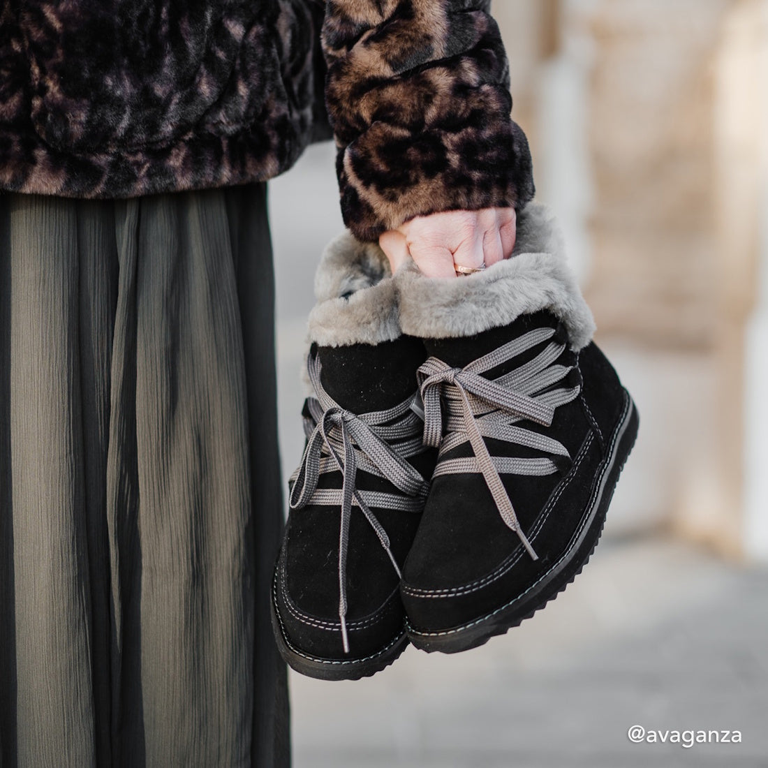 A person in a furry animal-print jacket and pleated pants holds Emu Womens Thorsby Black Sheepskin Ankle Boot Waterproof, featuring gray laces, arch support, and faux fur lining. The neutral background is slightly blurred., 9330071980507, 9330071980514, 9