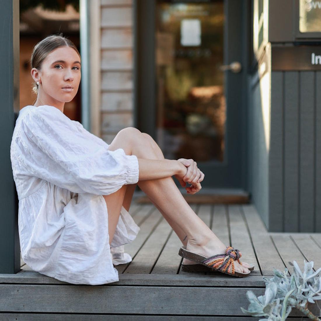 A person in a loose white dress sits on a wooden porch, gazing at the camera. They wear Emu Women's Zebedee Slide Sandals with a colorful plaited textile upper, and their light brown hair is tied back. A building entrance appears in the background., 93300