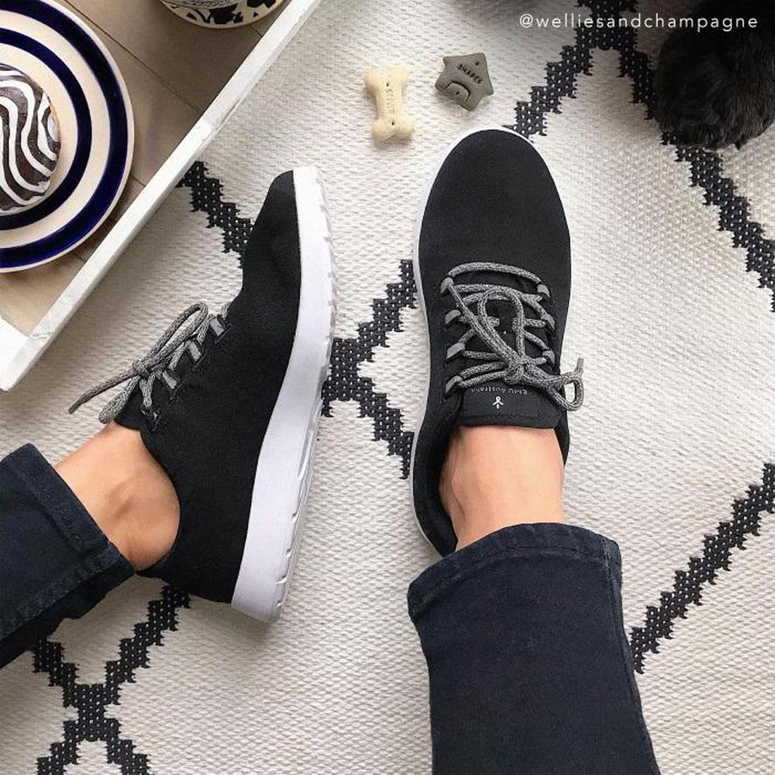 A person wearing Emu Barkly Black Wool Fabric Sneaker Lace Ups with white soles stands on a patterned rug, surrounded by dog toys and bowls that hint at a pet-friendly home., 9330071816479, 9330071816486, 9330071816493, 9330071816509, 9330071816516, 93300