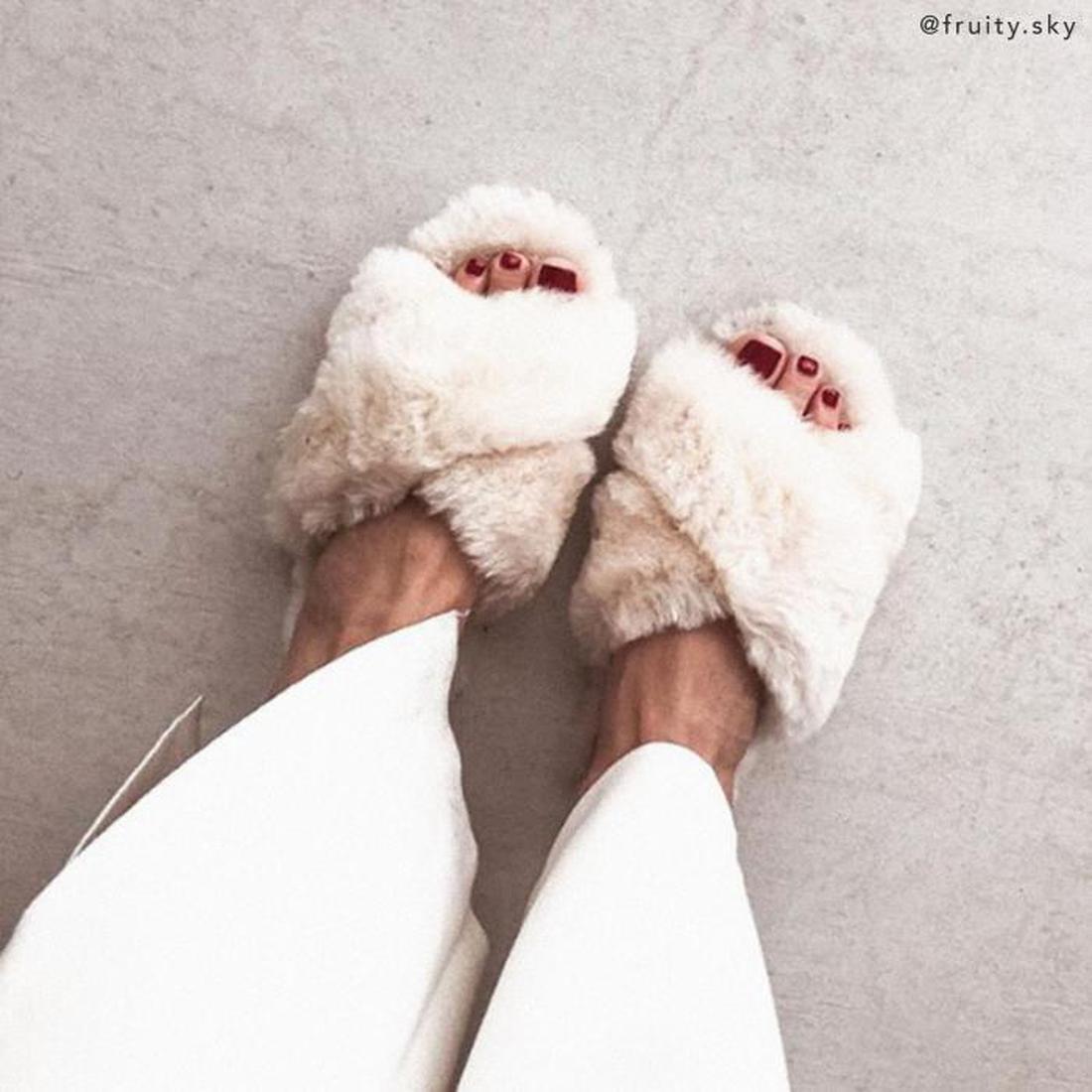 A person wears EMU Women's Mayberry Natural Luxurious Sheepskin Scuff Slippers with white pants, revealing red-painted toenails while standing on a light-colored floor., 9330071670002, 9330071670019, 9330071670026, 9330071670033, 9330071670040, 9330071670