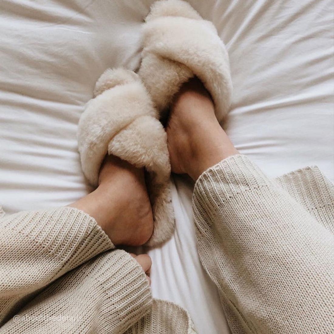 A person wearing EMU Women's Mayberry Natural Luxurious Sheepskin Scuff Slippers and matching knit pants is relaxing on a white bed with soft, wrinkled sheets., 9330071670002, 9330071670019, 9330071670026, 9330071670033, 9330071670040, 9330071670057
