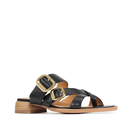 The EOS Teia Black Low Heeled Sandal Slide features black cashmere leather, two wide crisscross straps with large gold buckles, a low wooden block heel, and a tan sole for an elegant look.