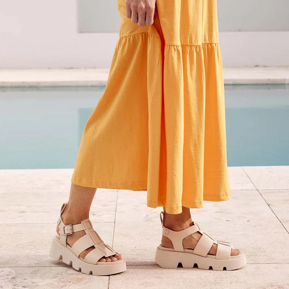 A person wearing a long yellow dress and EOS Womens Kailan Platform Chunky Leather Sandals in Bone stands on a light stone surface near a pool with calm blue water., 34434, 9321191131504, 9321191131511, 9321191131528, 9321191131535, 9321191131542, 9321191