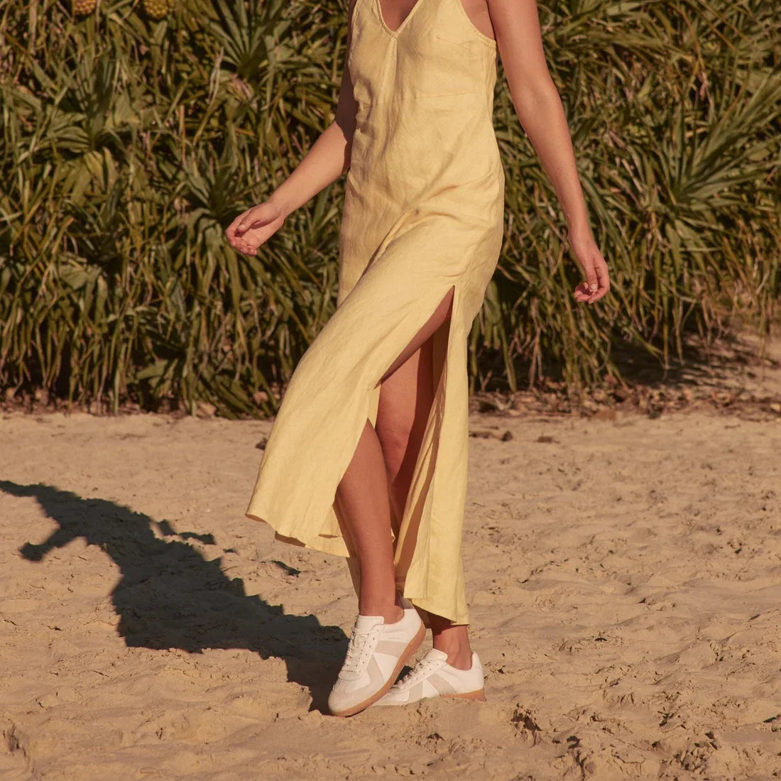 A person in a sleeveless yellow dress with a high slit walks barefoot on sand, holding EOS Melbaa White Leather Lace Up Trainers, with green plants behind them. Their head is not visible., 41691, 9321191224626, 9321191224633, 9321191224640, 9321191224657,