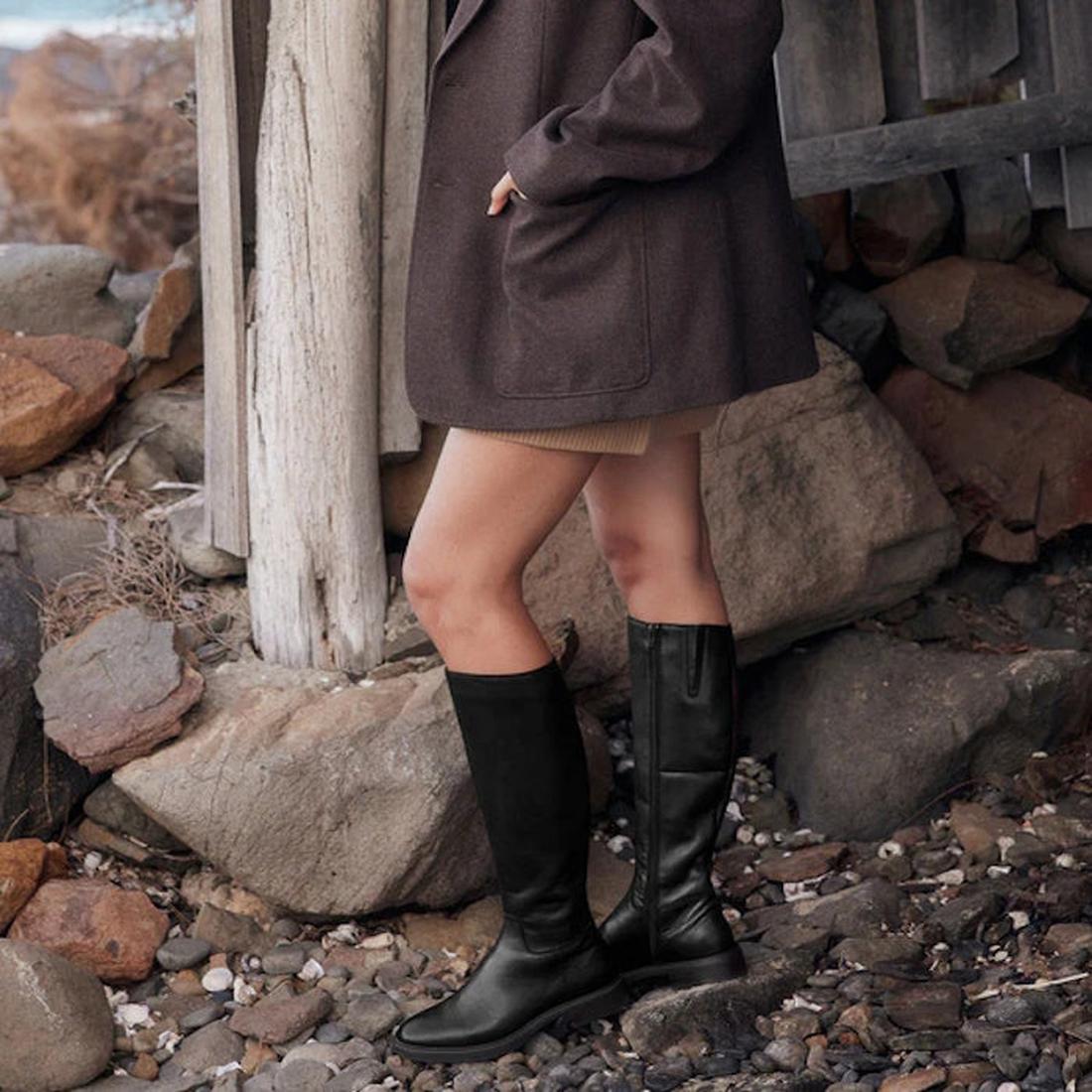 A person stands on rocky ground, wearing EOS Blanche Womens Black Leather High Boot Low Heel with a dark brown coat and tan skirt. Large rocks and wooden beams are in the background. The head and upper body are not visible., 9321191187808, 9321191187815, 
