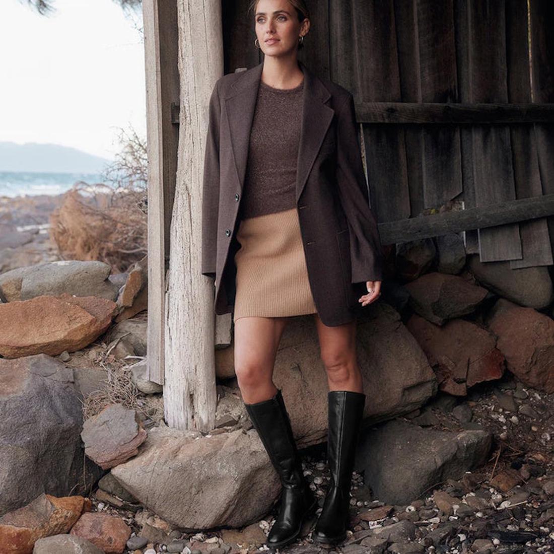 A woman stands outdoors by a rustic wooden structure, wearing the EOS Wyoming Black High Leather Women’s Heeled Boot, along with a brown blazer, brown sweater, and tan skirt. Rocky ground and distant water are seen in the background., 9321191201733, 93211