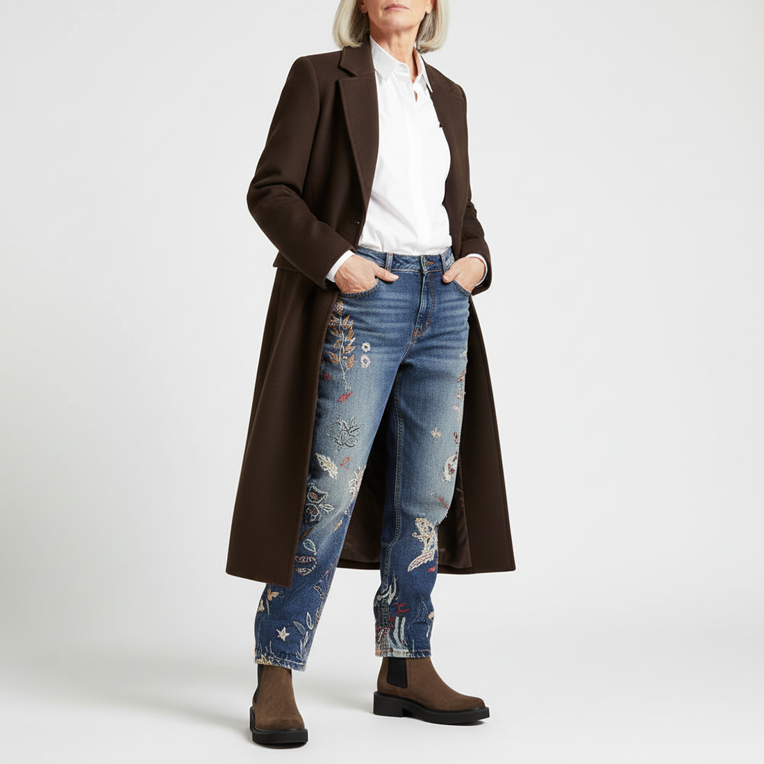 A person wearing EOS Womens Leilin Suede Kalamata Brown Ankle Pull On Boots, a long brown coat, white button-up shirt, and blue embroidered jeans stands with hands in jean pockets against a plain white background., 9321190037555, 9321190037562, 9321190037