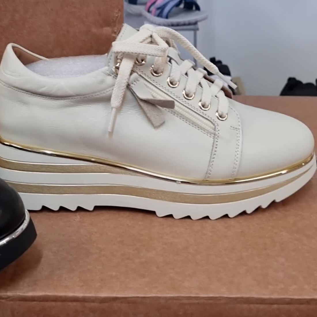 The Alfie & Evie Hosting Womens Leather Shoe in Cream/Gold, featuring a thick platform sole and gold accents, is displayed atop a brown box, with other footwear softly blurred in the background.