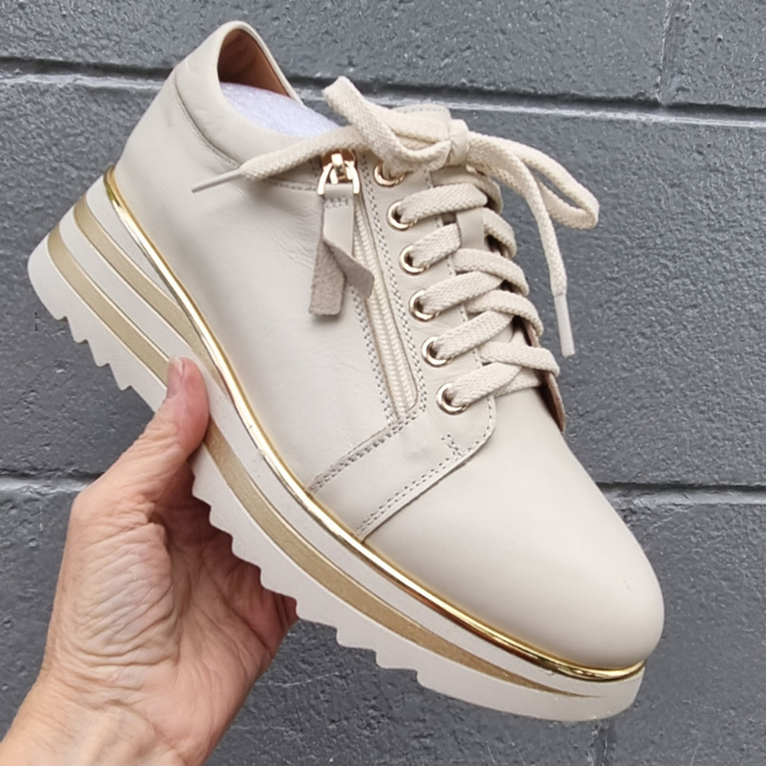 A hand holds the Alfie & Evie Hosting Womens Leather Shoe Platform in Cream/Gold, featuring a beige platform sole, gold accents, white laces, and a zip detail near the laces, set against a gray brick wall background.