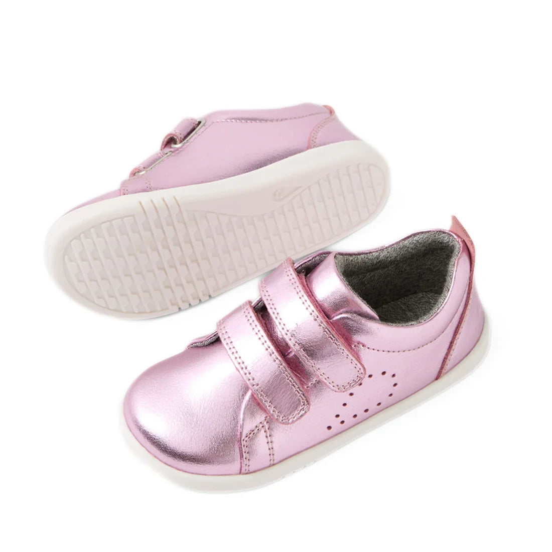 A pair of Bobux Iwalk Toddlers Grass Court Pink Metallic Leather Trainers, featuring shiny pink leather, white soles, and two Velcro straps, shown from both a side and bottom view—perfect trainers inspired by the classic Grass Court style., 9420074806888,