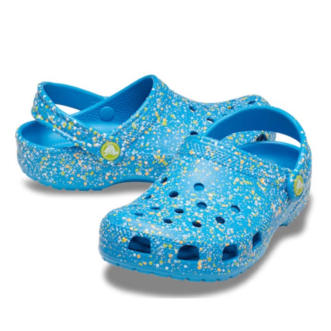 The Crocs Kids - Youths Classic Clog K Paint Splatter Print Blue Multi features blue clogs with yellow and white splatter patterns, ventilation holes, and a pivoting heel strap—ideal for kids' everyday comfort.