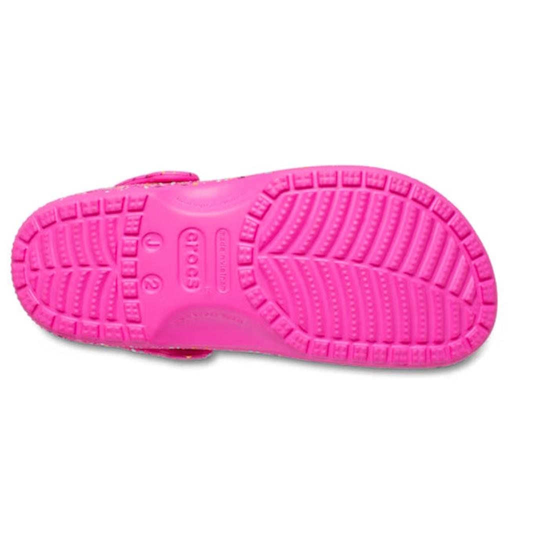 The Crocs Kids - Youths Classic Clog K Paint Splatter Print Bright Pink Multi is shown from the bottom, highlighting its textured rubber sole with patterns, grooves, and the Crocs logo embossed in the center., 198445210228, 198445210235, 198445210242, 198