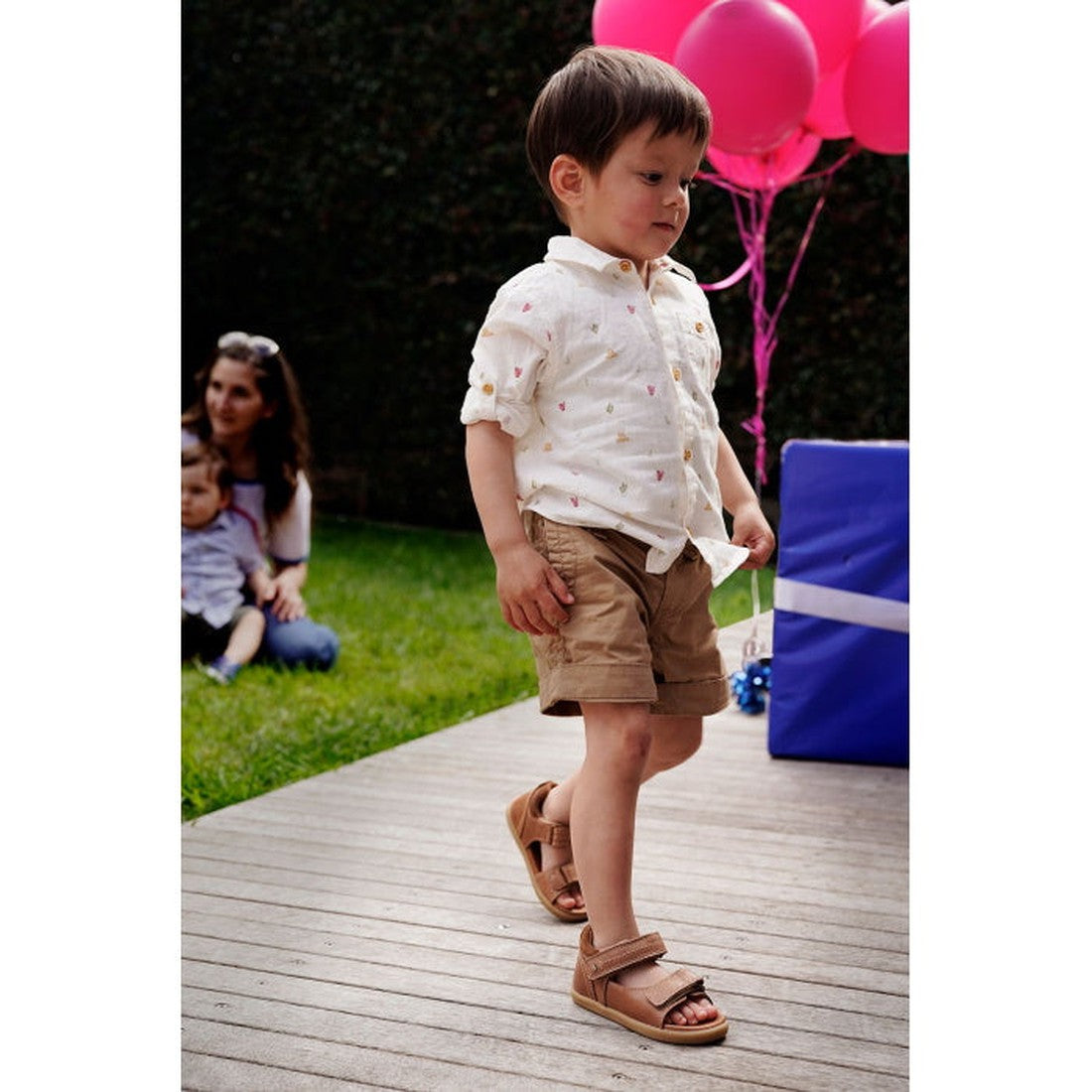 A young boy wearing a white shirt, tan shorts, and Bobux IWalk Driftwood Sandal Caramel Open Toe 633606A walks on a wooden deck. Pink balloons, a large blue gift box, and two people on the grass are in the background., 9420062048085, 9420062048078, 942006