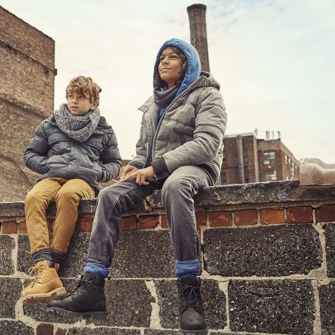 Two boys bundled up for winter relax outdoors, wearing Timberland Kids Black Icon 6-Inch Waterproof Boots with sturdy soles and laces.  ksnz