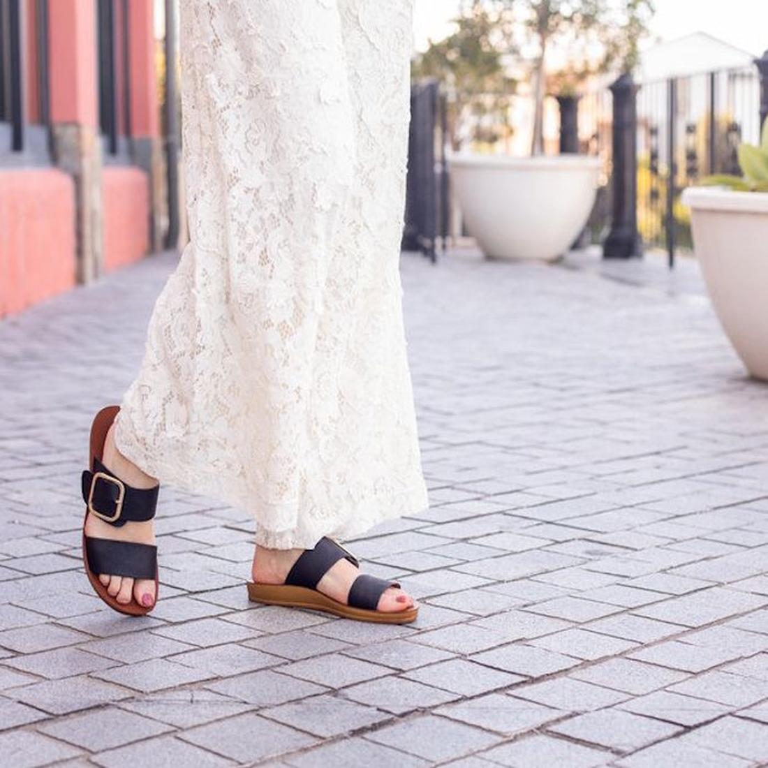 A person in a long white lace dress walks on a paved sidewalk lined with large planters, wearing Los Cabos Doti Black Light Flexible-Sandals., 9321191045450, 9321191045467, 9321191045474, 9321191045481, 9321191045498, 9321191045504, 9321191045511