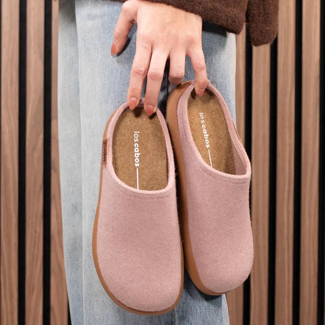 A person in blue jeans holds a pair of Los Cabos Felton Graphic Lavender Women's Felted Slipper House slippers, featuring pink felt and cushioned brown soles, against a backdrop of vertical wooden panels., 9321190006025, 9321190006032, 9321190006049, 9321