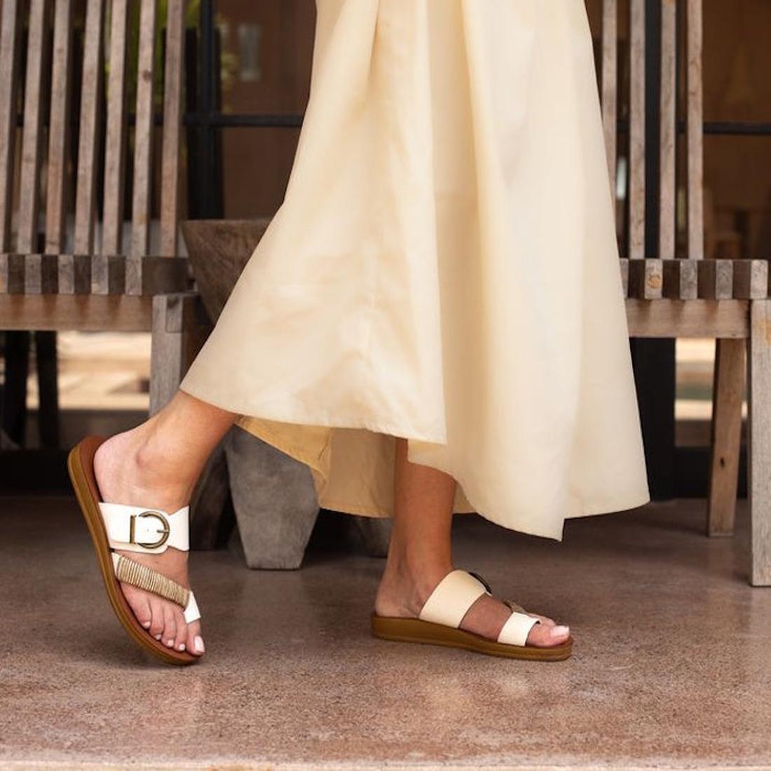 A person wearing a long cream skirt stands on a polished stone floor, showcasing two different off-white summer sandals with brown soles from Los Carbos—one of them being the stylish Bria Off-White Light Flexible Sandal Jandal. A wooden bench and a stone 