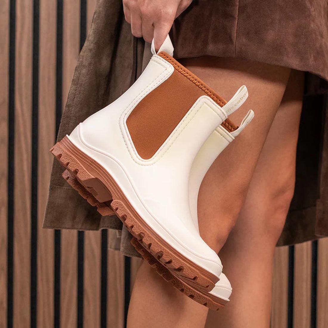 A person in a brown skirt holds the Los Cabos Amelia Cream Ankle Rain Boot Waterproof—trendy chunky Chelsea-style rain boots with rugged soles—standing before a vertical wooden slat wall., 9321190004977, 9321190004984, 9321190004991, 9321190005004, 932119