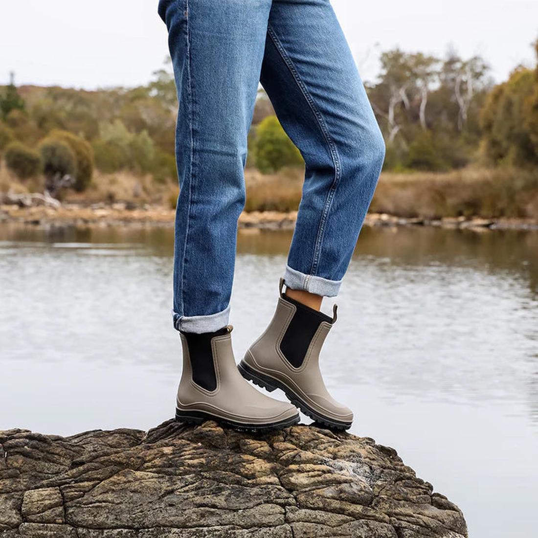 A person in rolled-up blue jeans and Los Cabos Amelia Khaki Ankle Rain Boot Waterproof stands on a rock by a calm lake, with trees and shrubs in the background., 50795, 9321191166711, 9321191166728, 9321191166735, 9321191166742, 9321191166759, 93211911667