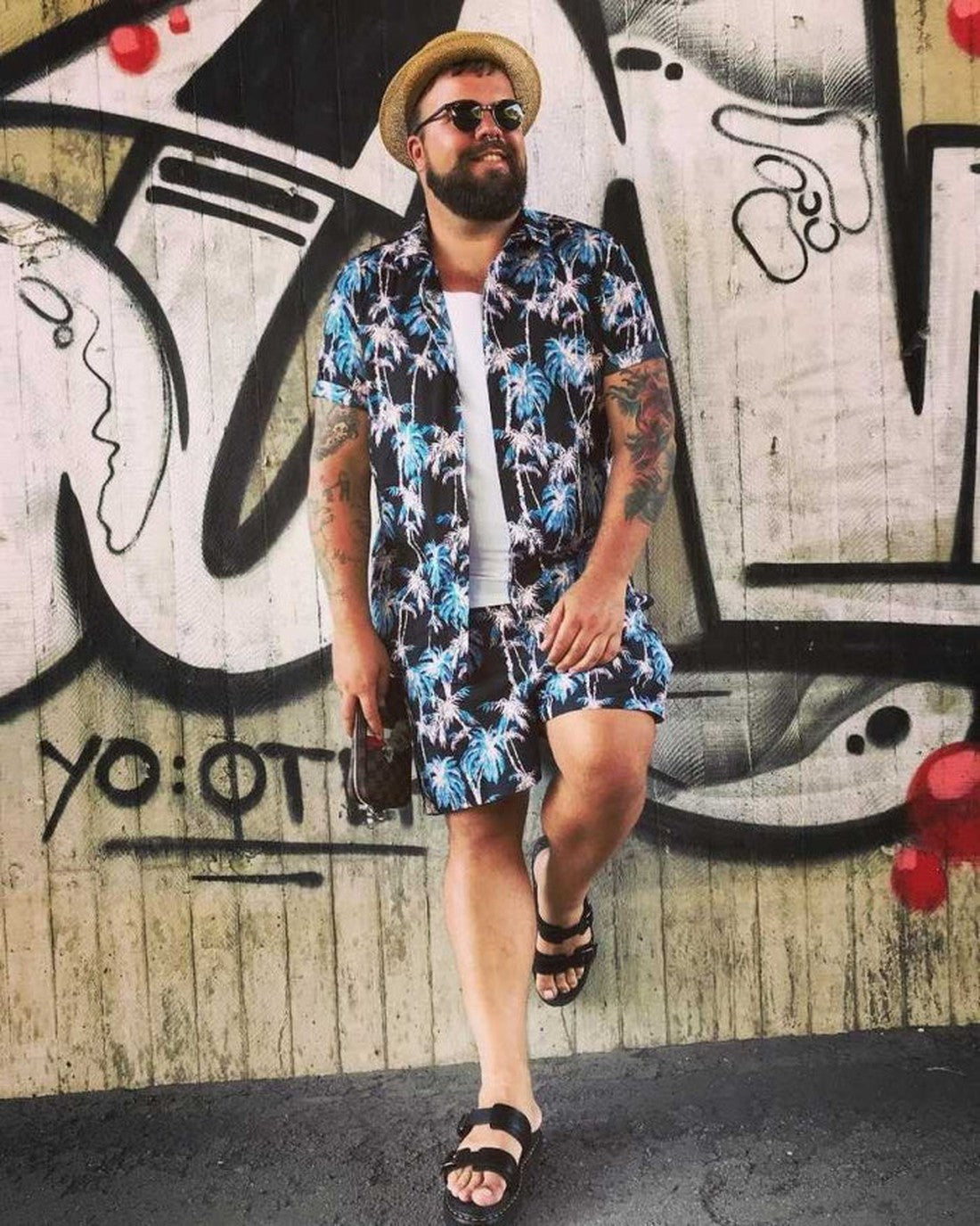 A bearded man in sunglasses, a straw hat, and a palm tree print outfit leans against a graffiti wall, smiling with his phone. His tattooed arms stand out as he wears stylish Dr Martens Black Myles Bar Brando Sandals with adjustable buckle straps., 1906651