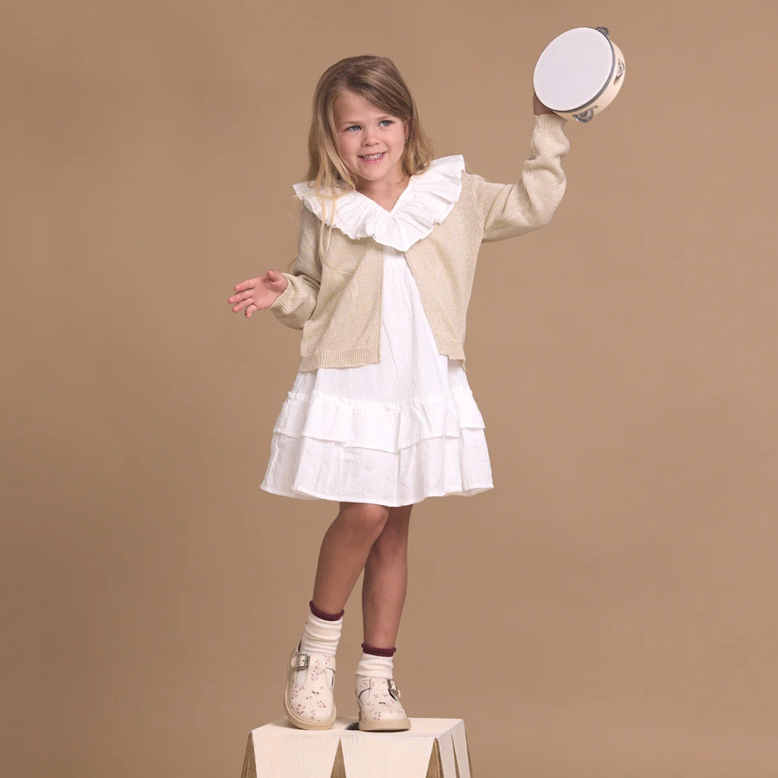 A young girl in a white dress and beige cardigan stands on a wooden block, smiling and holding a tambourine above her head, wearing Pretty Brave Girls Maddison Winter Bloom Pink Leather Mary Jane T Bar shoes against a plain tan background., 9420075362840,