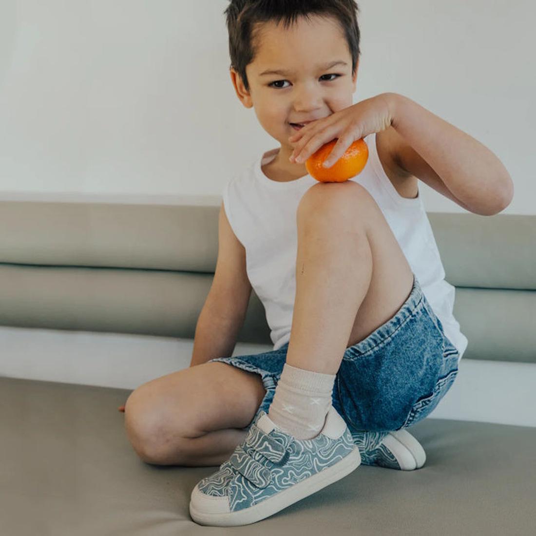 A young boy in a white tank top and denim shorts sits on a couch, smiling and holding an orange near his mouth. He wears Pretty Brave Otto Organic Canvas Trainer With Leather Toe Buffer Blue Ripple and beige socks, with one knee bent up., 9420075314337, 9