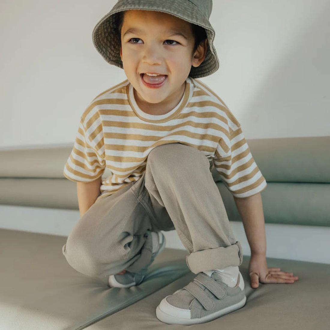 A young child in a striped beige and white t-shirt, light pants, a bucket hat, and Pretty Brave Otto Organic Canvas Trainer With Leather Toe Buffer Khaki smiles while crouching on a sofa., 9420075314597, 9420075314580, 9420075314573, 9420075314566, 942007