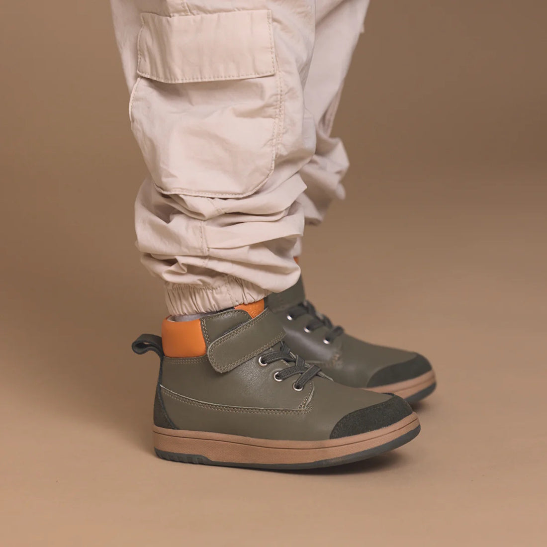 A child wears light beige cargo pants and Pretty Brave Hi Top Boot Corey Olive kids leather boots with black soles and an orange heel accent, standing against a tan background., 50474, 50475, 9420075364370, 9420075364363, 9420075364356, 9420075364349, 942