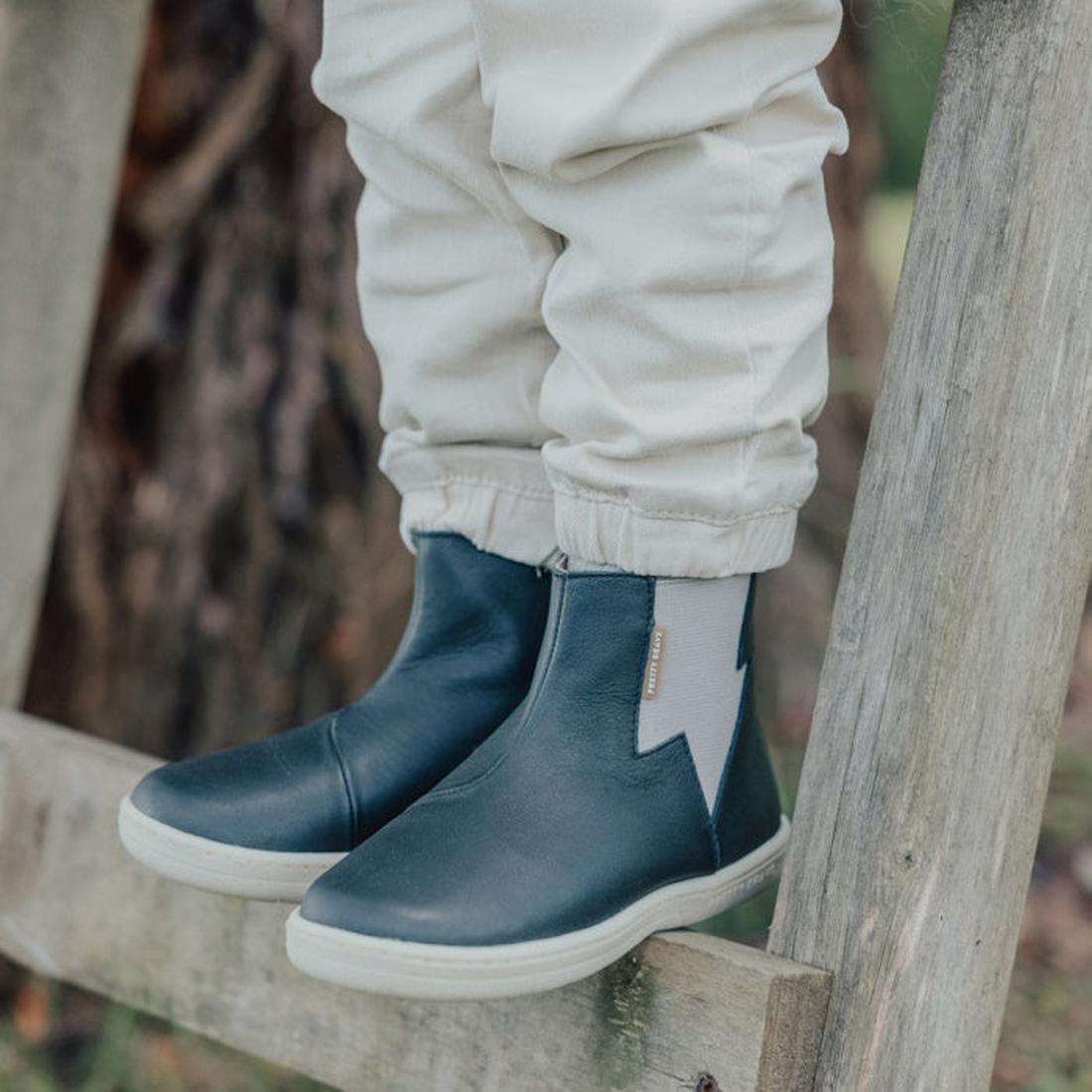 A child in light-colored pants and Pretty Brave Electric Leather Boot - Jet Black, featuring a lightning bolt design and APMA approval, stands outdoors on a wooden fence or ladder., 35922, 9420075327986, 35924, 35925, 9420075327955, 9420075327948, 9420075