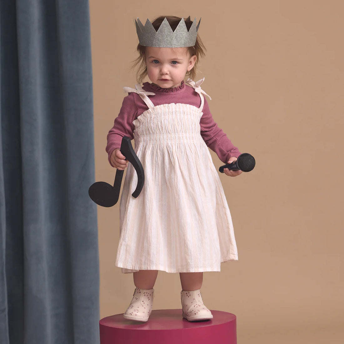 A young child in a paper crown and pink top stands on a round platform holding large musical note props, wearing Pretty Brave Charlie Leather Boot First Walkers Winter Bloom Pink, with a neutral background and blue curtain to the side., 9420075361881, 942