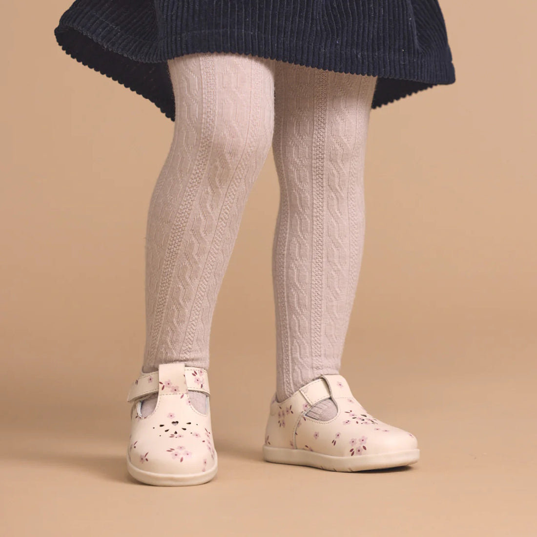 A young child wears a navy blue skirt, white cable-knit tights, and Pretty Brave Miley First Walker Mary Jane T Bar Leather Winter Bloom Pink shoes, standing against a beige background., 9420075361980, 9420075361973, 9420075361966, 9420075361959