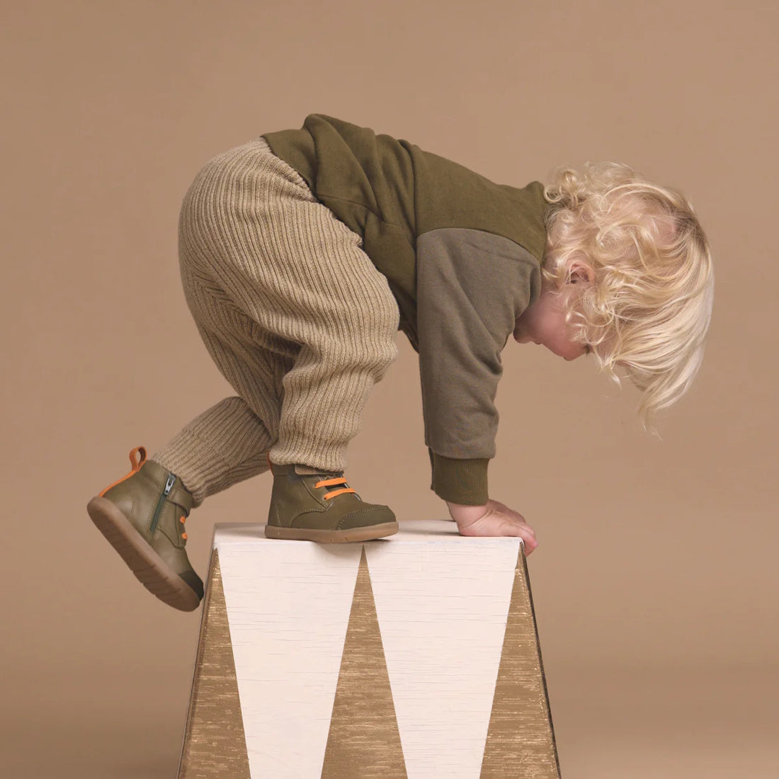 A young child with blonde curly hair wears Pretty Brave Jessie First Walker Hi Top Leather Olive Green shoes while climbing a white and gold triangular block—podiatrist-approved style showcased against a beige background., 9420075361782, 9420075361775, 94