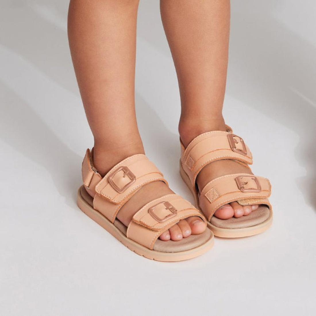 A child stands on a white surface wearing Pretty Brave Alex Leather Sandal Coral (sizes 29-33, clearance, not returnable) featuring adjustable velcro straps and buckles., 9421036357554, 9421036357561, 9421036357578, 9421036357585, 9421036357592