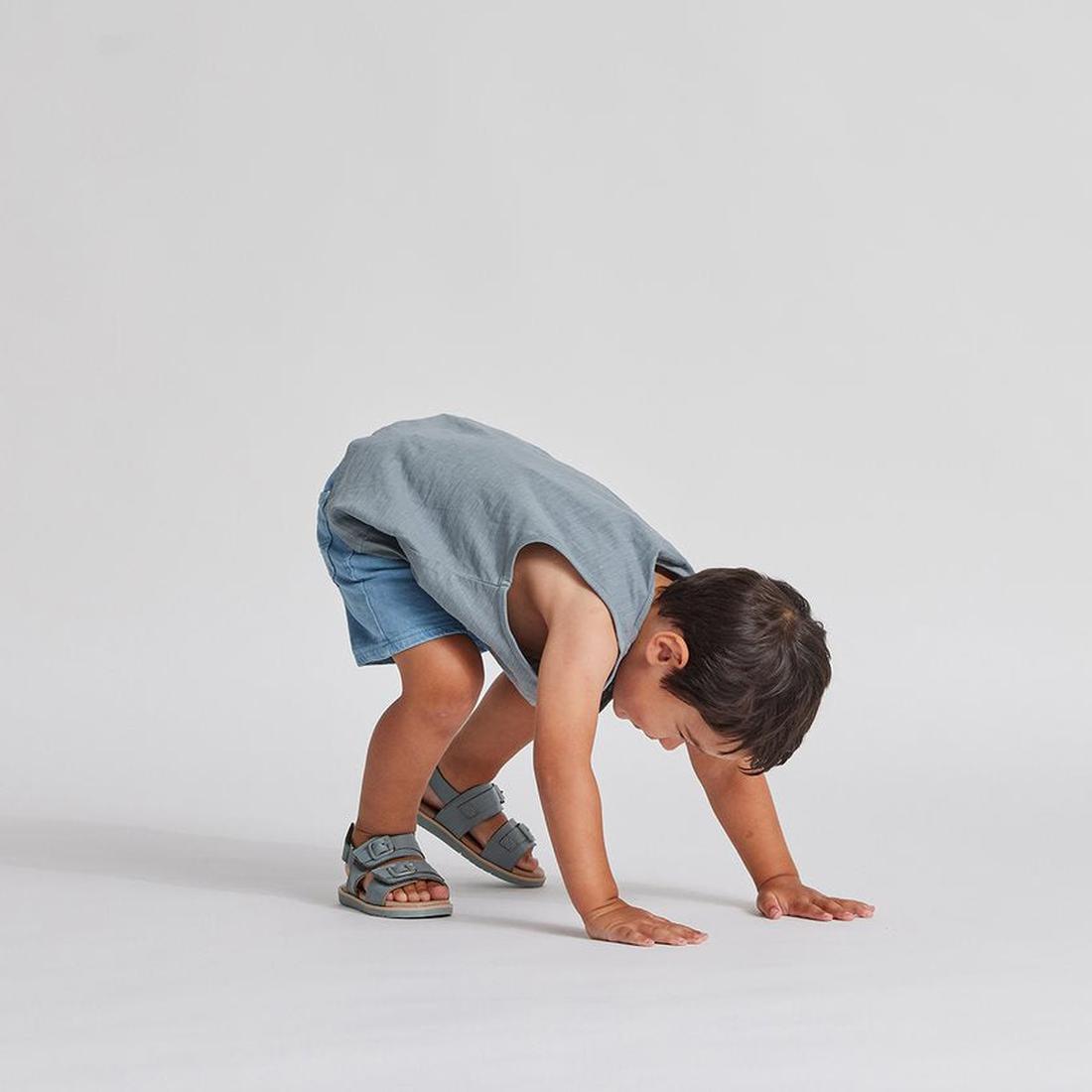 A young child in a gray sleeveless shirt, denim shorts, and Pretty Brave Alex Leather Sandal Seaform Blue (sizes 29-33, CLEARANCE NOT RETURNABLE) bends over with hands on the ground against a plain light background., 9421036357684, 9421036357691, 94210363