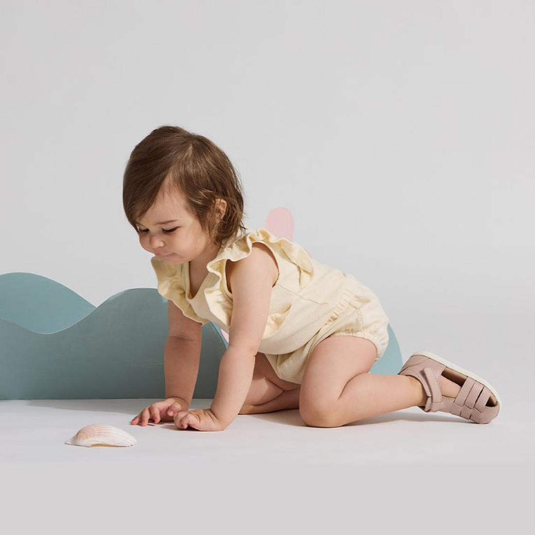 A toddler wearing a light yellow outfit and Pretty Braver Billie First Walker Sandal Closed Back Blush crawls on a white surface, reaching for a seashell, with a pastel blue wave-shaped prop behind them against a plain background., 9420075334458, 94200753