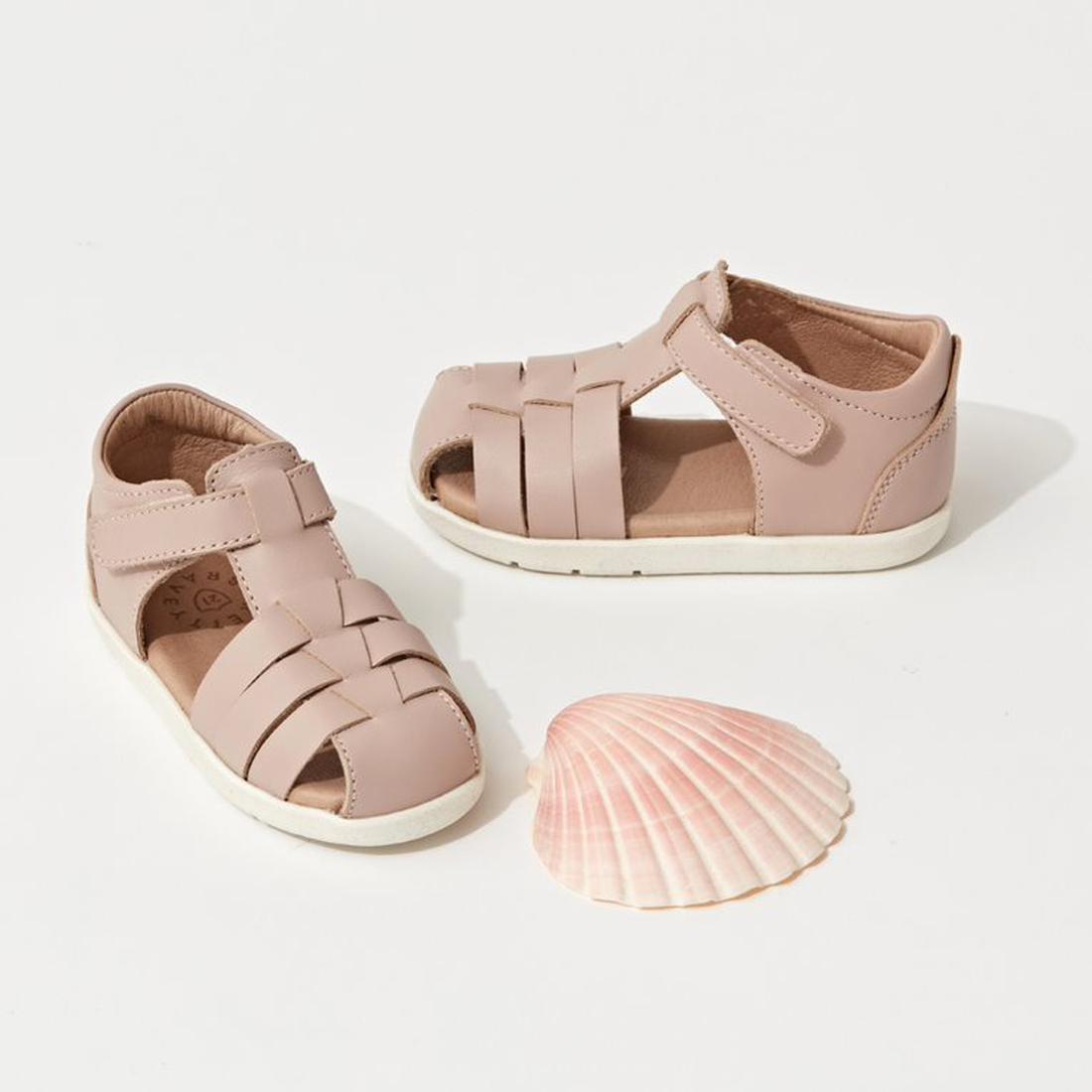 The Pretty Braver Billie First Walker Sandal Closed Back Blush, featuring light pink color with white soles, is displayed beside a pink seashell on a white background—ideal as an early walker sandal for toddlers., 9420075334458, 9420075334441, 94210363571