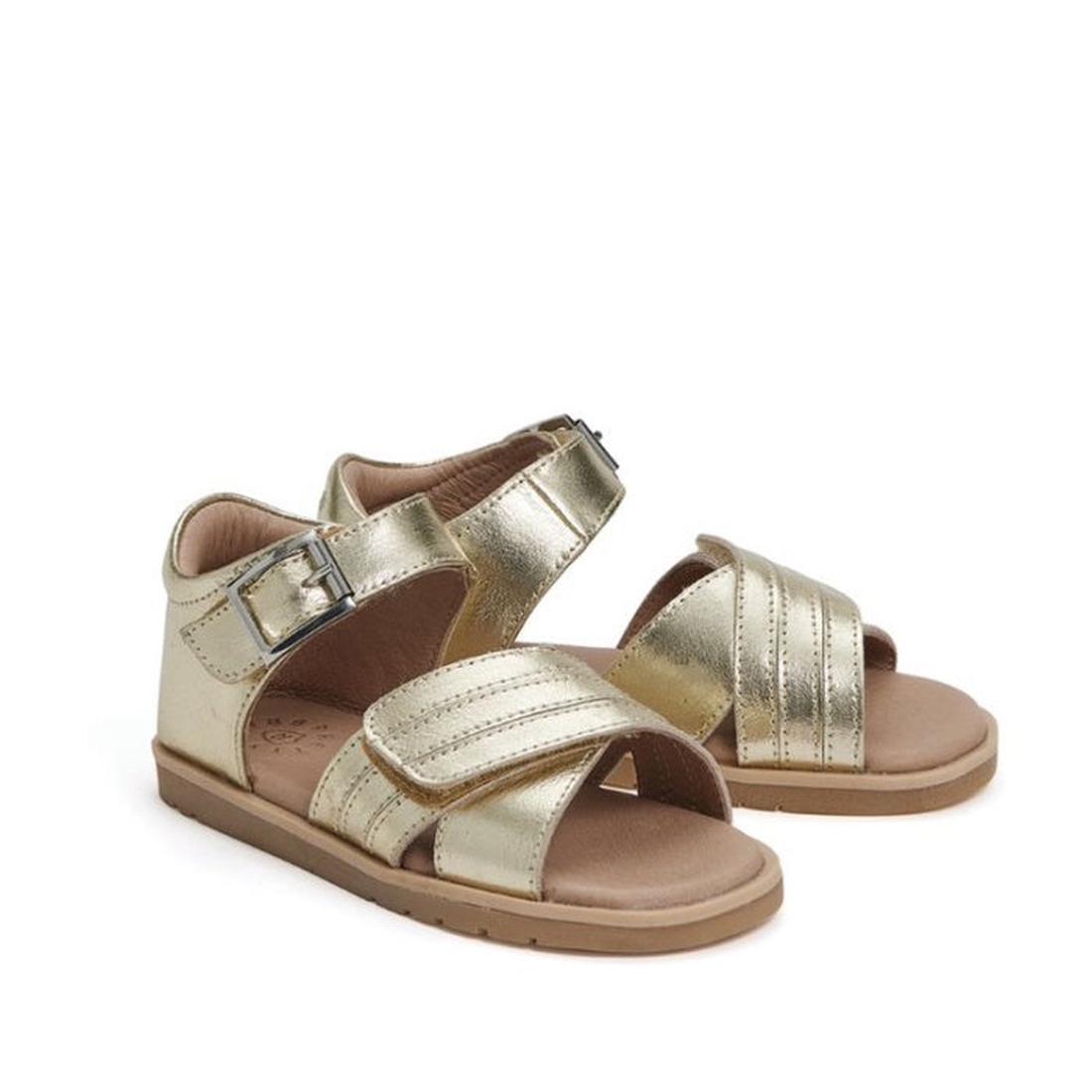 The Pretty Brave Georgia Gold Leather Sandal 23-28 is a girls’ sandal with open toes and heels, wide crisscross straps, side buckles, a cushioned footbed for comfort, and flat tan soles.