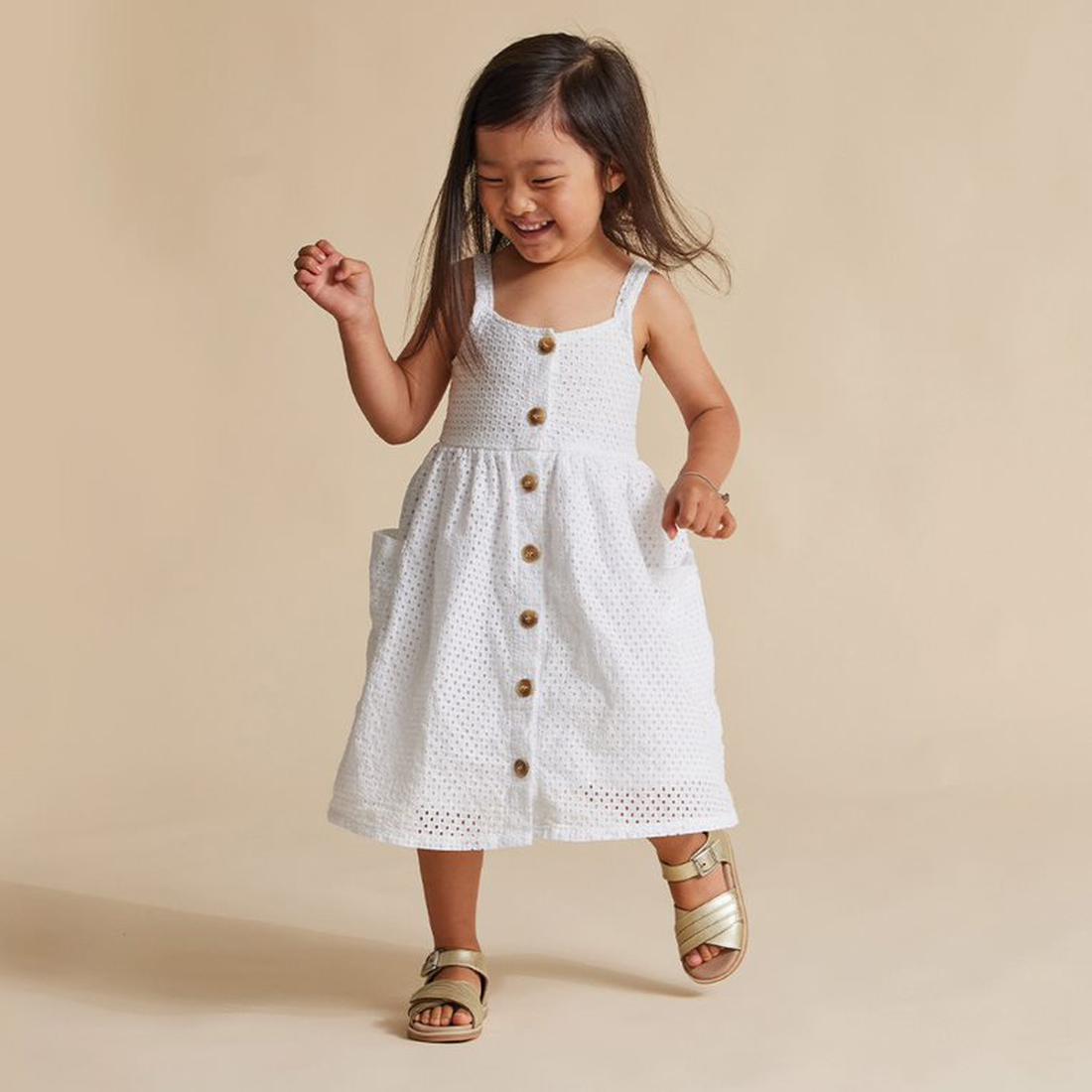 A young girl with long dark hair smiles and plays, wearing a sleeveless white dress and Pretty Brave Georgia Gold Leather Sandals (sizes 23-28) with a cushioned footbed, standing against a plain beige background., 25302, 9421036358278, 9421036358285, 9421