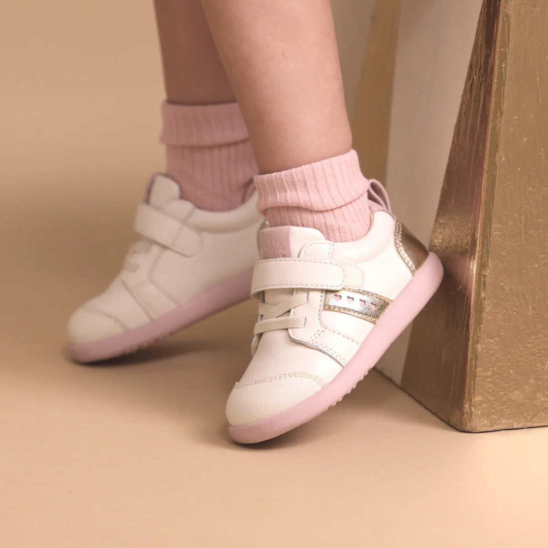 A child wearing light pink socks and Pretty Brave SID Leather Trainer First Walker Off White Stone Pink rests their feet against a gold-painted block on a beige background., 9420075362963, 9420075362956, 9420075362949, 9420075362932
