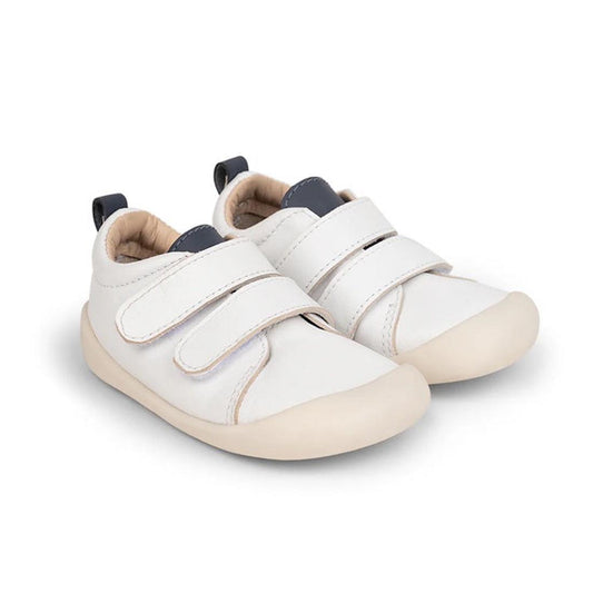 The Pretty Brave First Walker Toto Leather Trainer in White Indigo is a pair of flexible baby shoes designed to support little feet on their first adventures, featuring white leather uppers, beige soles, two hook-and-loop straps, and navy pull tabs at the