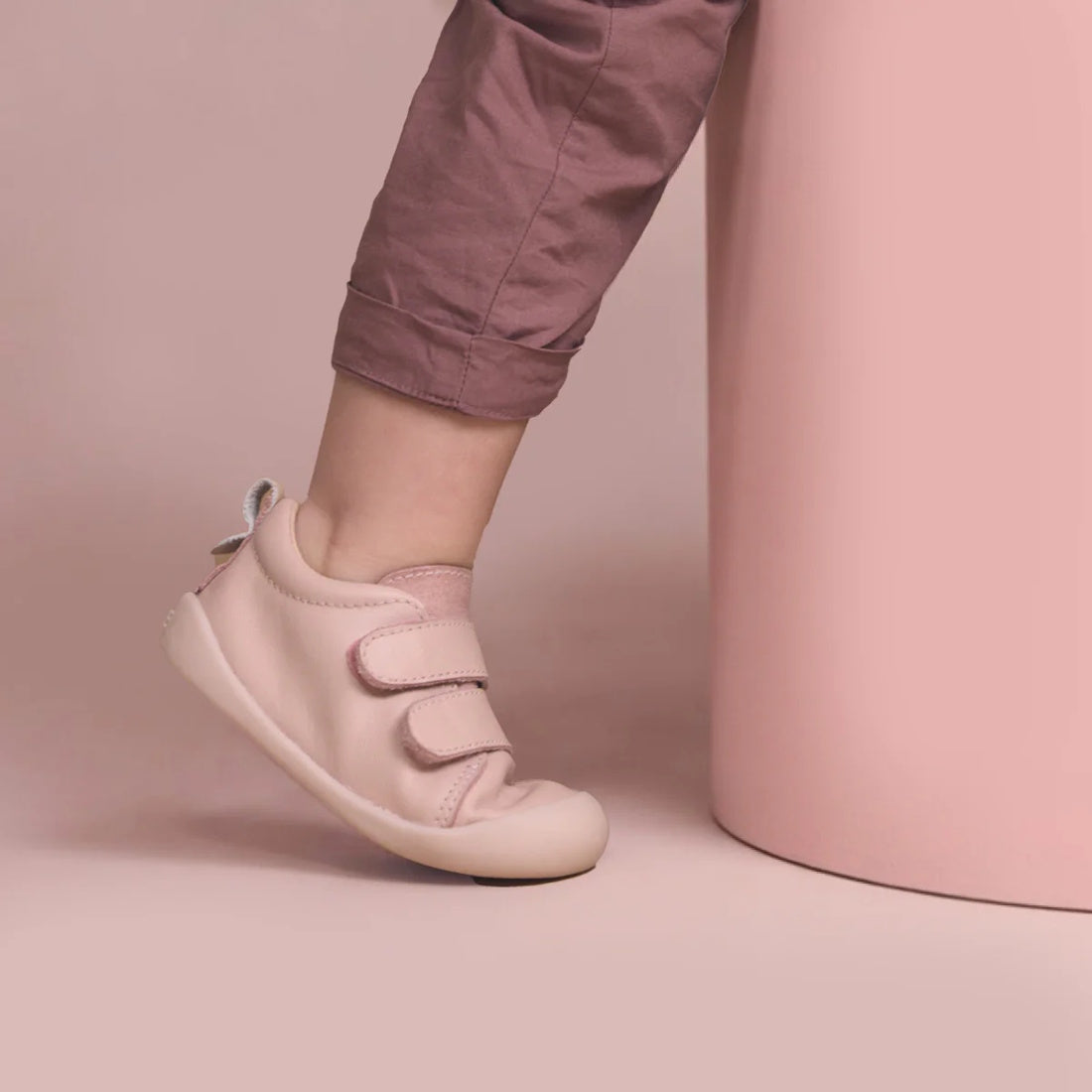 A toddler stands with one foot slightly lifted, wearing mauve pants and Pretty Brave First Walker Toto Leather Trainer shoes in Dusky Pink, beside a light pink cylinder against a soft pink background., 9420075366329, 9420075366312, 9420075366305, 50976