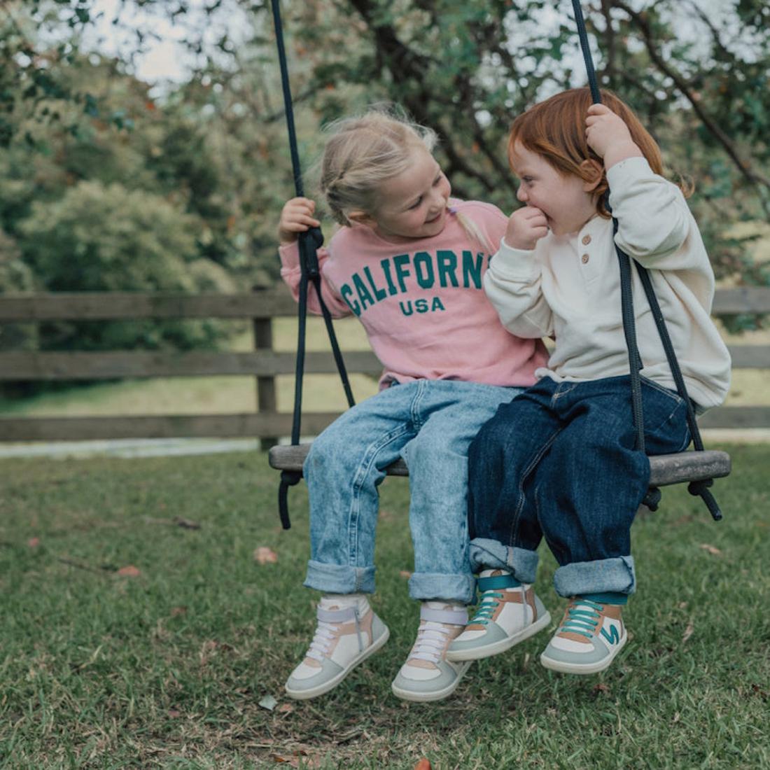 Two young children smile at each other on a wooden outdoor swing. One wears a pink California USA sweatshirt and Pretty Brave Hi Top Boot Forest Mix sneakers; the other is in a white top. Trees and a wooden fence are in the background., 35902, 35903, 3590