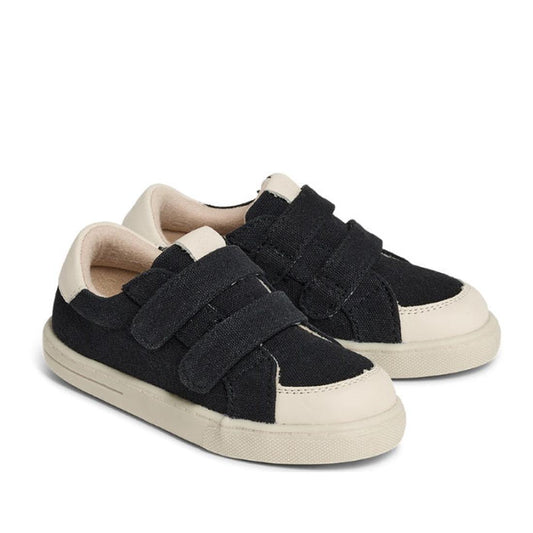A pair of Pretty Brave Otto Organic Canvas Trainers in black with leather toe buffer, white soles, bamboo cotton lining, and two velcro straps per shoe, displayed on a white background.