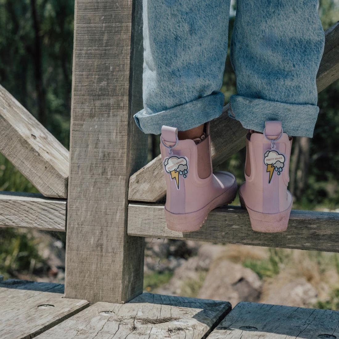 A person wearing rolled-up blue jeans and Pretty Brave Puddle Boot Ankle Rainboot Blush Pink stands on a wooden fence, with green trees and rocks in the background., 9420075328525, 9420075328518, 9420075328501, 9420075328495, 9420075328488, 9420075328471,