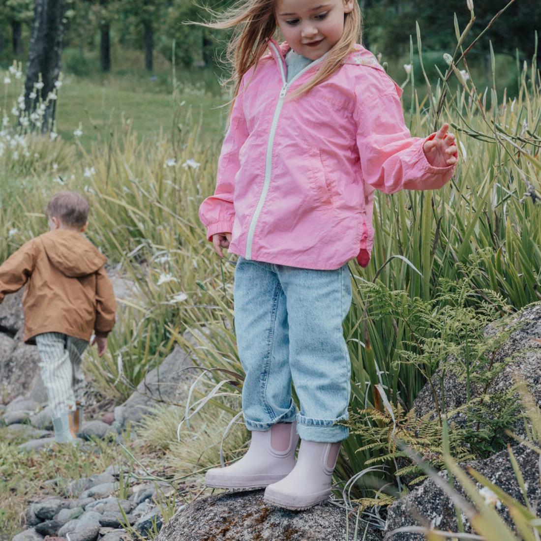 A young girl in a pink jacket and jeans stands on a rock in a grassy area, wearing Pretty Brave Puddle Boot Ankle Rainboot Blush Pink, while another child in a brown jacket walks away. Both kids seem to be exploring nature., 9420075328525, 9420075328518, 