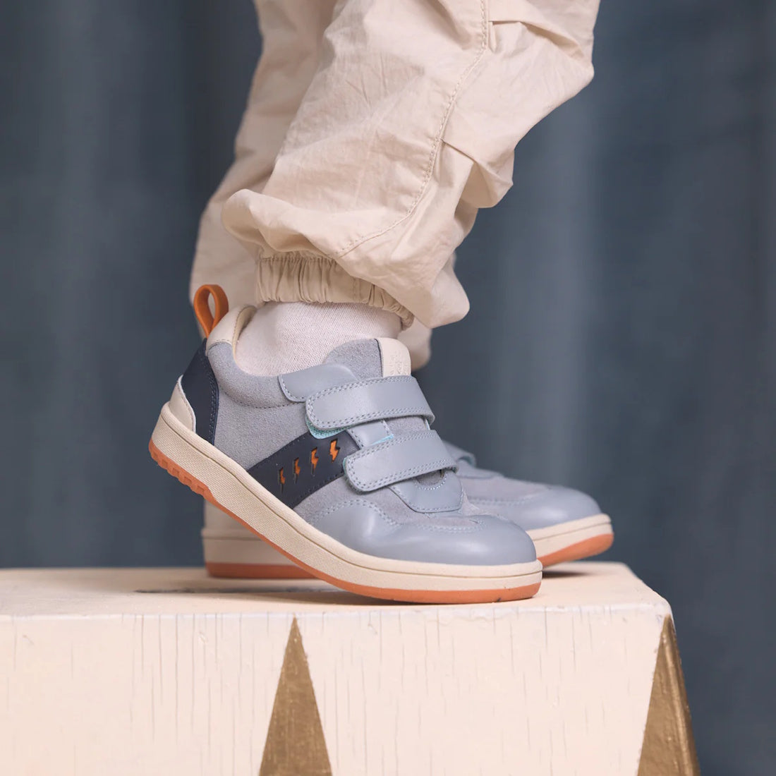 A child in light-colored pants and white socks stands on a white wooden surface, wearing Pretty Brave River Bolts Leather Trainer Blue Grey sneakers with podiatrist approval, Velcro straps, and orange accents., 9420075363984, 9420075363977, 9420075363960,