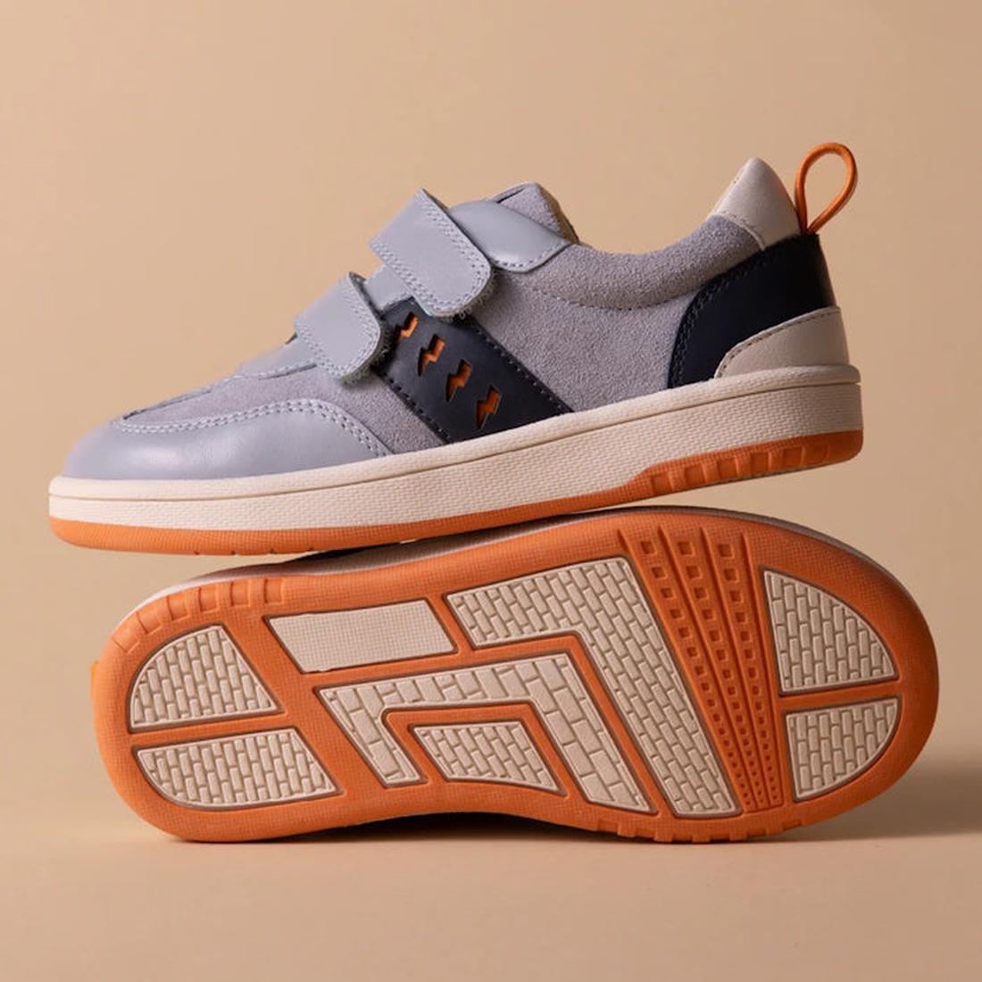The Pretty Brave River Bolts Leather Trainer Blue Grey is a kids' sporty shoe in light gray and navy with orange accents and white soles. One shoe stands upright, while the other lies on its side to display the textured orange and white sole., 94200753639